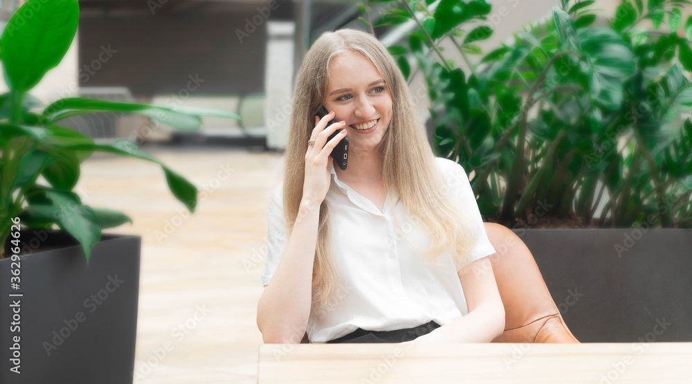 Portrait of a cheerful young caucasian blond business woman sitting on the table, talking at phone and smiling.Concept of successful young women in a corporation. Girl in a white shirt. 