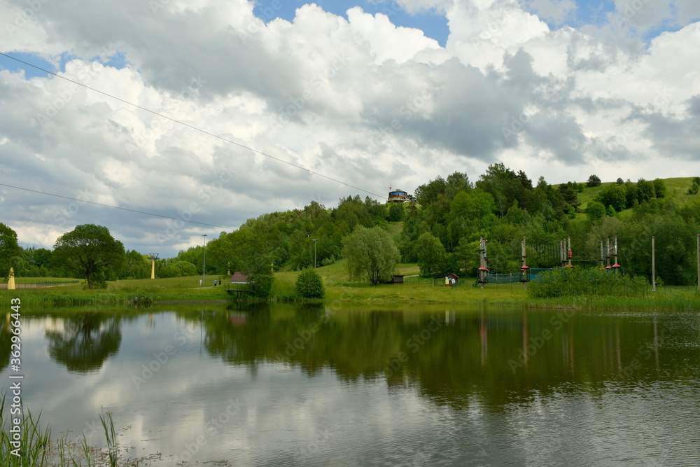 View of a tall hill with a tower on top overgrown with massive trees and shrub located next to a small lake or pond reflecting the cloudy summer sky seen during a hike on a Polish countryside