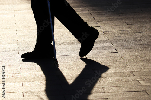 Silhouette of limping man walking with a cane, shadow on pavement. Concept for disability, old age, blind person, dramatic life