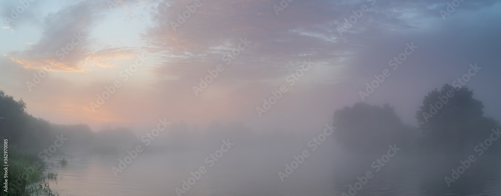 Tranquil morning landscape panorama at sunrise. Heavy fog over the river. Dawn illuminates and makes the clouds colorful.