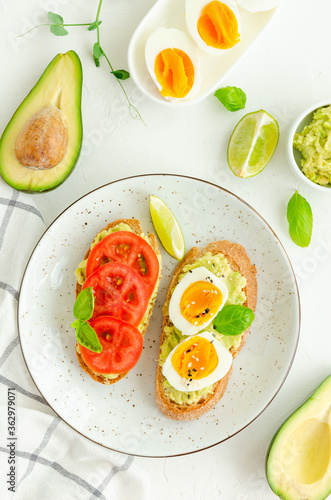 Toasts with mashed avocado, boiled eggs, fresh tomato and basil on a white concrete background. Healthy food. Vertical, copy space.