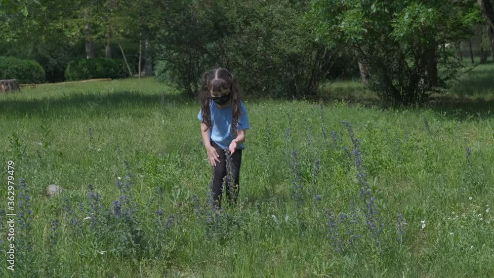 Alone child in coronavirus time. A little girl in a protective mask collects flowers on a green lawn in the park. Alone child in coronavirus time. A little girl in a protective mask collects flowers on a green lawn in the park.