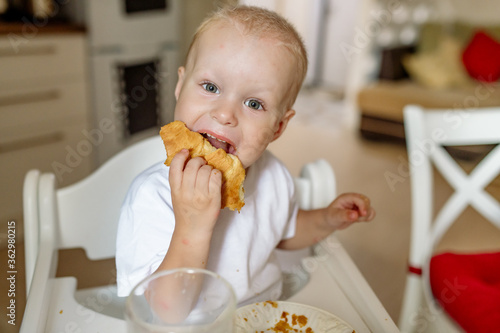 Cute 2 years old boy eating a delicious homemade bun while sitting in a highchair in the kitchen