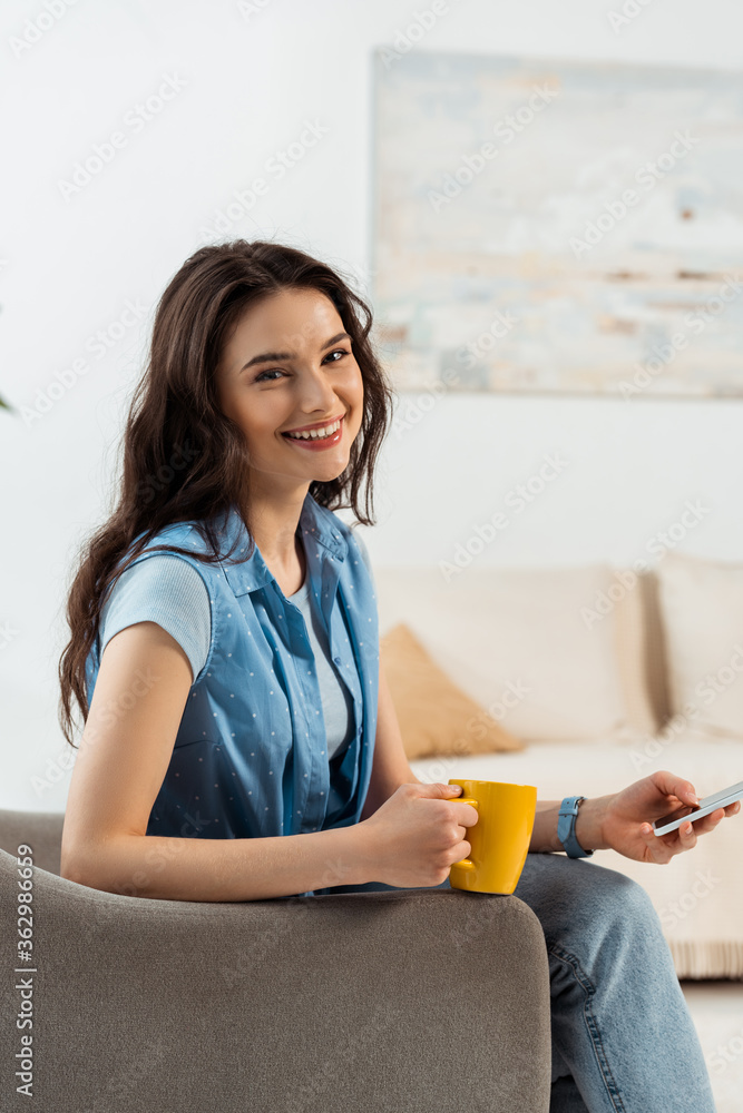 Beautiful woman smiling at camera while using smartphone and holding cup of coffee in armchair Beautiful woman smiling at camera while using smartphone and holding cup of coffee in armchair