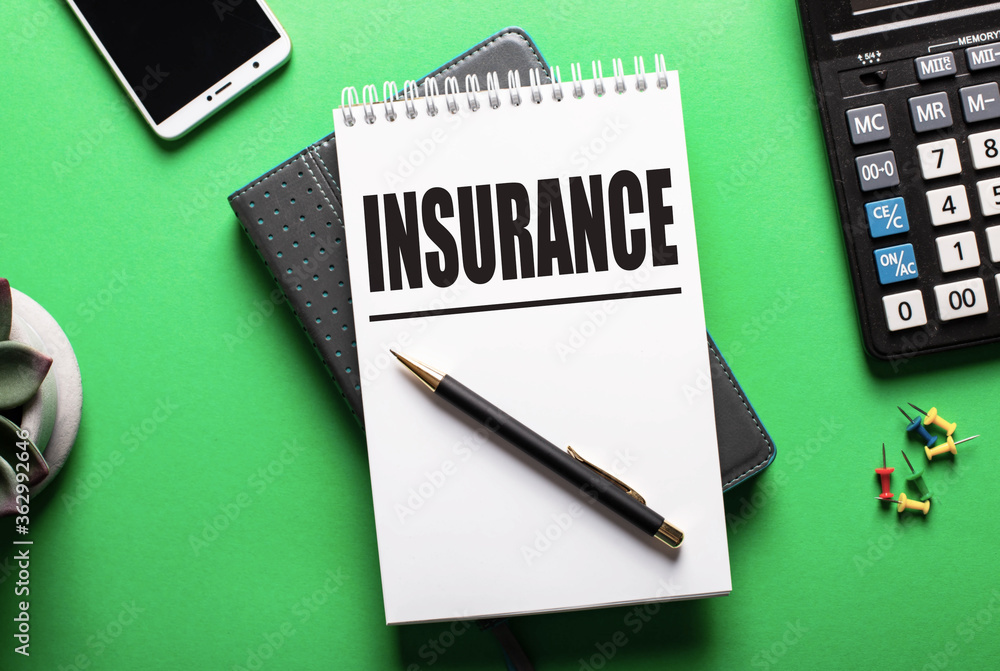 The word INSURANCE is written in a white notebook on a green table near a calculator and a phone. Insurance concept The word INSURANCE is written in a white notebook on a green table near a calculator and a phone. Insurance concept