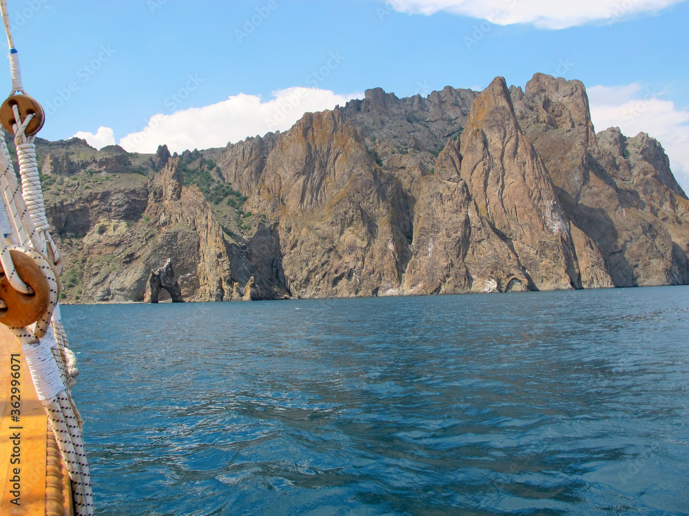 A picturesque panorama of the impregnable cliffs of the extinct volcano Karadag from the side of a pleasure yacht against a bright blue sky with white clouds and blue sea
