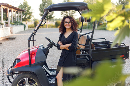 Cheerful woman with curly hair in black dress holding in hands a laptope, poses near vehicle and smiling. Small bussines content. photo