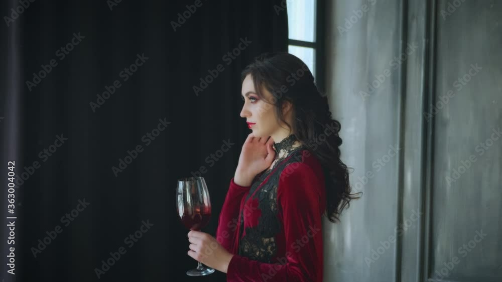 Young beautiful sexy fairytale mystical vampire queen. Woman lady holds glass bloody wine in hand. Brunette black long wavy hair. Red velvet masquerade vintage dress. Dark interior gothic room, window Young beautiful sexy fairytale mystical vampire queen. Woman lady holds glass bloody wine in hand. Brunette black long wavy hair. Red velvet masquerade vintage dress. Dark interior gothic room, window