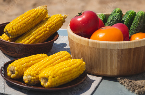 boiled corn on the cob on an earthenware plate. fresh tomatoes and cucumbers in a wooden plate. natural food.