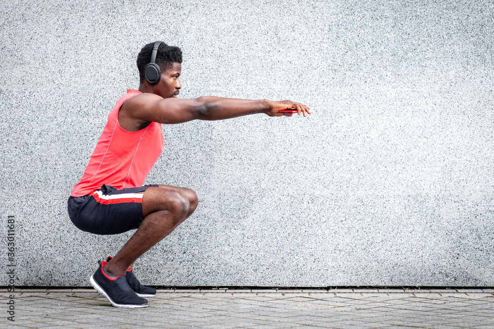 african american sports guy working out with headphones outdoors against wall, athletic man crouches and does physical exercises