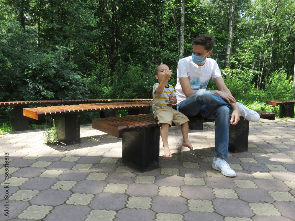 Fototapeta premium Father and son in the park, sitting on a bench and drinking some water. Man in a medical mask on a walk. 