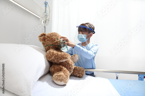 child in the hospital puts oxygen mask on teddy bear on bed, wearing protective visor and surgical mask, corona virus covid 19 protection concept.