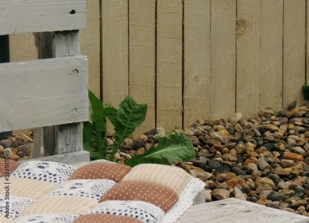 Soft pillows with floral print on a white wooden bench on the street, in the background a green dandelion and stones. Soft pillows with floral print on a white wooden bench on the street, in the background a green dandelion and stones.