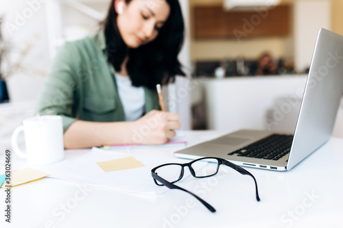 Desktop. Glasses. In the background  in defocus  a young student girl writes information in her notebook during online learning at home.