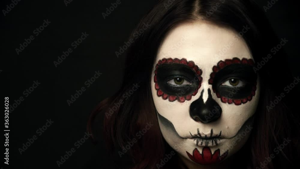 Girl with sugar skull makeup opens green eyes and her pupils get smaller. Portrait of woman on black background watching in camera. Theme of santa muerte makeup and emotions. Girl with sugar skull makeup opens green eyes and her pupils get smaller. Portrait of woman on black background watching in camera. Theme of santa muerte makeup and emotions.