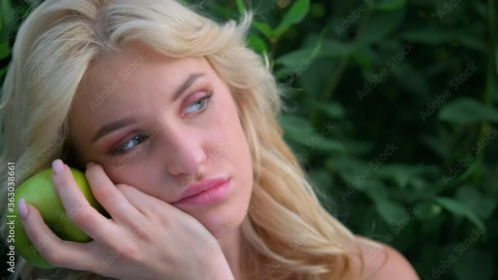 Portrait of a young beautiful woman with curly blond hair against a park and green trees. Beautiful face, blue eyes and luscious lips. The girl laughs playfully and holds a green apple in her hands. Portrait of a young beautiful woman with curly blond hair against a park and green trees. Beautiful face, blue eyes and luscious lips. The girl laughs playfully and holds a green apple in her hands.