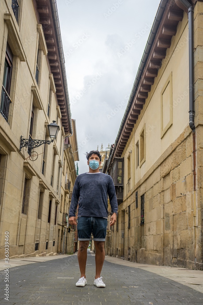 Vertical shot of a middle-aged Hispanic man with a medical mask on walking in between the buildings