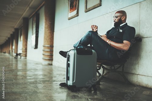 A mature African bald bearded businessman is waiting for a train to start his business trip, looks at the clock while sitting on a wooden bench at the platform of a railway station with a bag near him