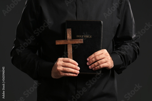 Young priest with Bible and cross on dark background