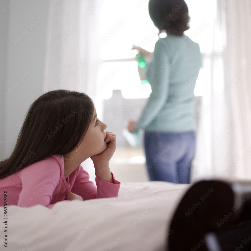 Girl daydreaming on the bed while her mother is cleaning the window Girl daydreaming on the bed while her mother is cleaning the window