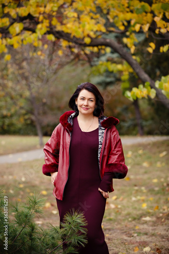 Fototapeta Naklejka Na Ścianę i Meble -  Autumn mood. Beautiful middle-aged woman weared in red jacket is smiling and walking in a park on background of beautifull autumn leaves. Fall coming. Autumn story. Yellow leaves in park. 