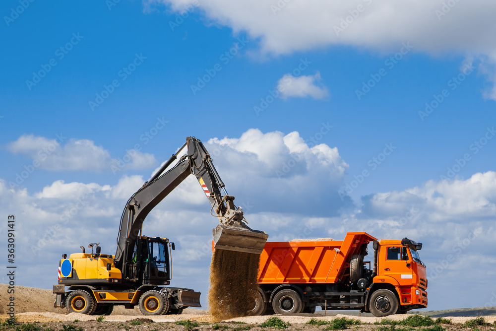 A yellow backhoe loader loads the earth into a truck during the construction of a road against a blue sky with clouds. A yellow backhoe loader loads the earth into a truck during the construction of a road against a blue sky with clouds.
