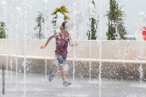 6 years old boy in a red T-shirt and gray shorts runs through the city fountains on a sunny summer day. Happy carefree childhood, lifestyle, freedom. Copy space.