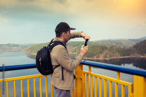 A man takes a photo of a river with a mobile phone. The tourist stays on the bridge