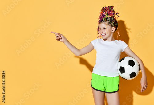 Laughing girl with colorful dreadlocks hairstyle in t-shirt, shorts stands with soccer ball in hand pointing at copy space photo