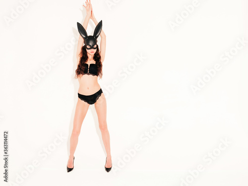 Beautiful sexy woman wearing carnival black mask of  rabbit.Hot blonde girl posing near white wall in studio. Seductive model in nice lingerie