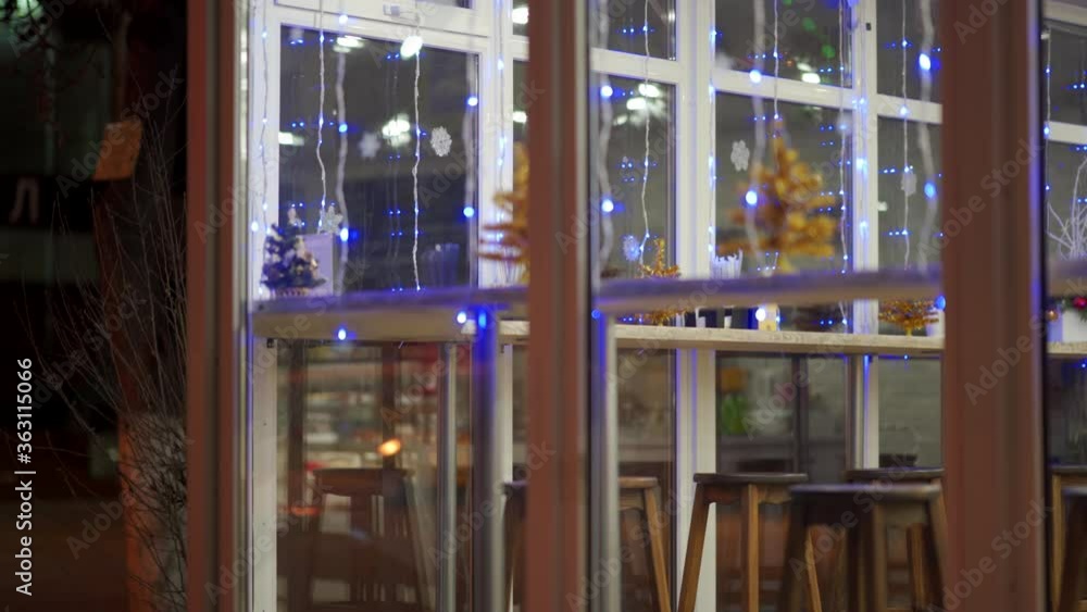 Coffee shop window display with festive lighting at dusk, window of cafe in the evening decorated with Christmas garlands, small Christmas trees on window counter. Coffee shop window display with festive lighting at dusk, window of cafe in the evening decorated with Christmas garlands, small Christmas trees on window counter.