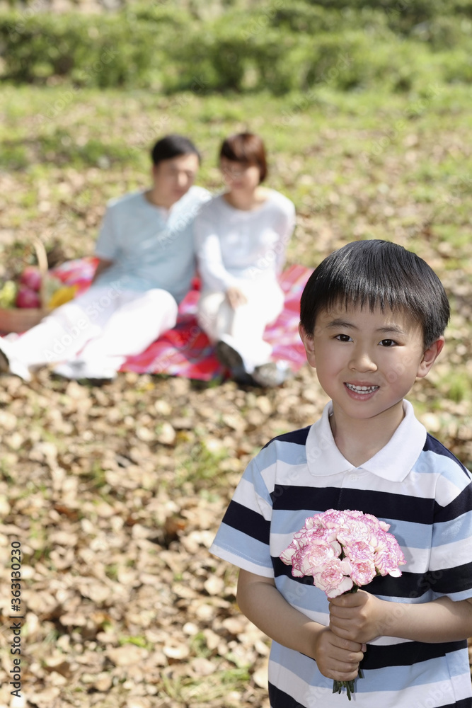 Boy holding a bunch of flowers, man and woman having a picnic in the background