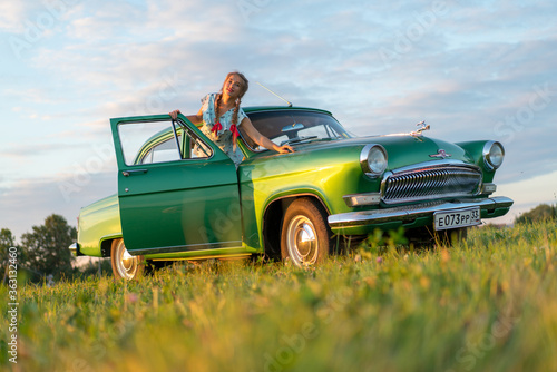 Beautiful Russian girl in stylistics with red ribbons in her hair in a field with a Russian church and the famous Russian car   Volga  of green color at sunset