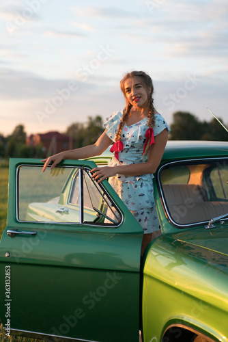 Beautiful Russian girl in stylistics with red ribbons in her hair in a field with a Russian church and the famous Russian car: "Volga" of green color at sunset