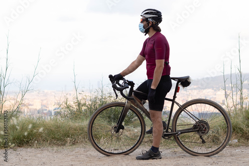  Male cyclist with a gravel bike observing the landscape with a medical mask. Concept: sport and coronavirus