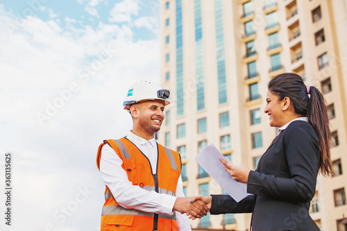 Indian engineer shaking hands with business woman over the office building on background