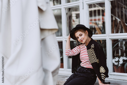 Konstfotografi Stylish young woman blonde in a hussar jacket, black hat in the city near the window of the restaurant