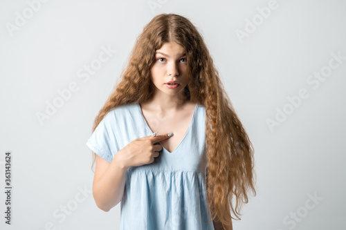 Me?! Portrait of indignant girl with curly long hair startled by offensive words, points at herself with for finger, opens mouth in surprisement, has dissatisfied expression, wears casual clothes