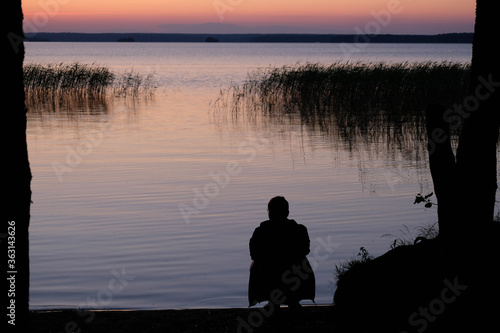  Sunset lake blurry silhouette of a young man looking at the lake  beautiful seascape with sunset. Outdoor summer lake view with dark silhouette. Man looks at the sunset lake. Scenic landscape. 