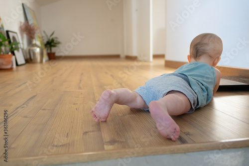 Little baby lying on belly on wooden floor with barefoot. Back view of adorable red-haired infant crawling at home. Childhood and infancy concept