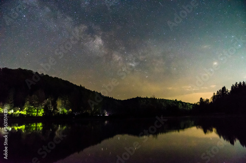 Germany, Slowly moving stars of milky way galaxy over swabian alb forest tree silhouette and reflecting in water of a lake at night