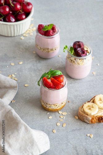 Homemade natural fruit yogurt with granola, strawberries, raspberries and cherries in glass jars on gray background. Healthy breakfast set.