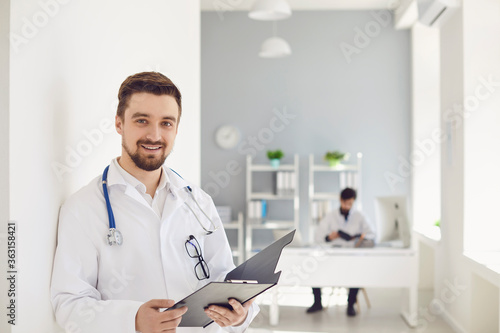 A male doctor with a stethoscope smiling a clipboard while standing in a clinic.