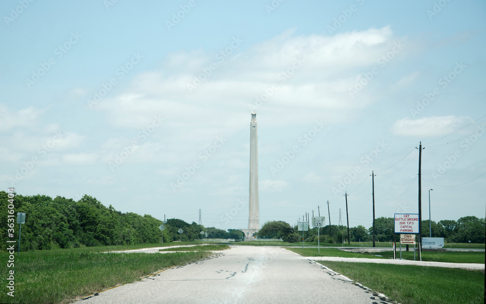 A view down the entrance road leading to the San Jacinto Monument. The tower is in the distance on a sunny day with a blue sky. A view down the entrance road leading to the San Jacinto Monument. The tower is in the distance on a sunny day with a blue sky.