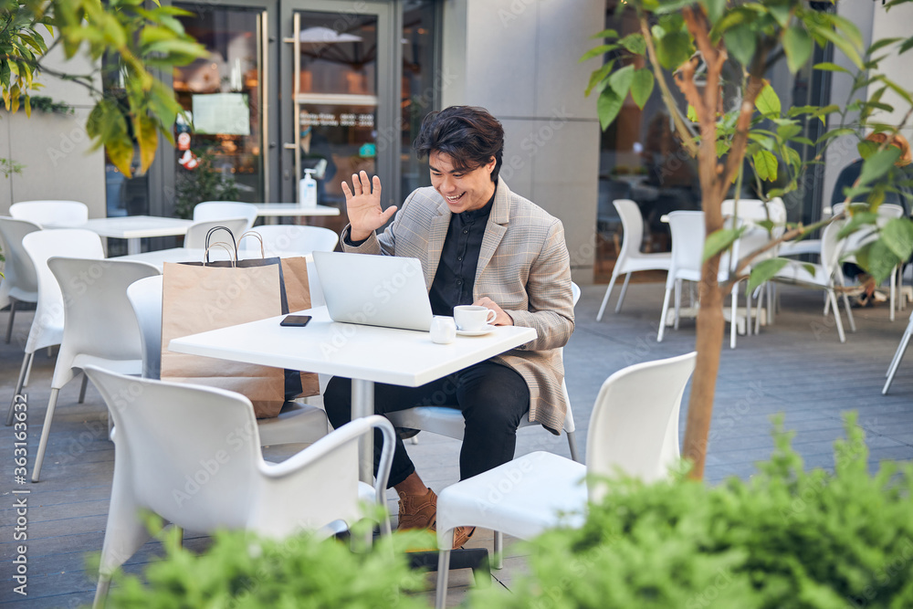 Cheerful young man chatting with friend online in outdoor cafe Cheerful young man chatting with friend online in outdoor cafe