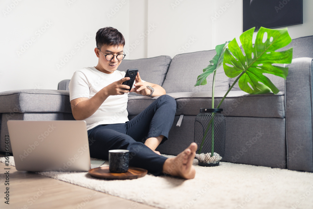 Young Asian man chatting with smartphone after working by using laptop computer. A man relaxing and sitting on floor in private room. Work from home concept