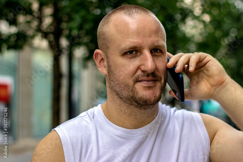 Joyful handsome sport man making phone call while standing on the street. Bearded man dressing in a white shirt using mobile.Young male in casual clothes standing outside and making call on smartphone