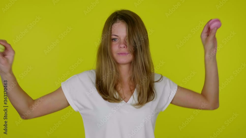 Funny woman in a white T-shirt is dancing with pink and yellow macaroons in hands on a yellow background with copy space. Place for text or product. 4K slow motion footage Funny woman in a white T-shirt is dancing with pink and yellow macaroons in hands on a yellow background with copy space. Place for text or product. 4K slow motion footage