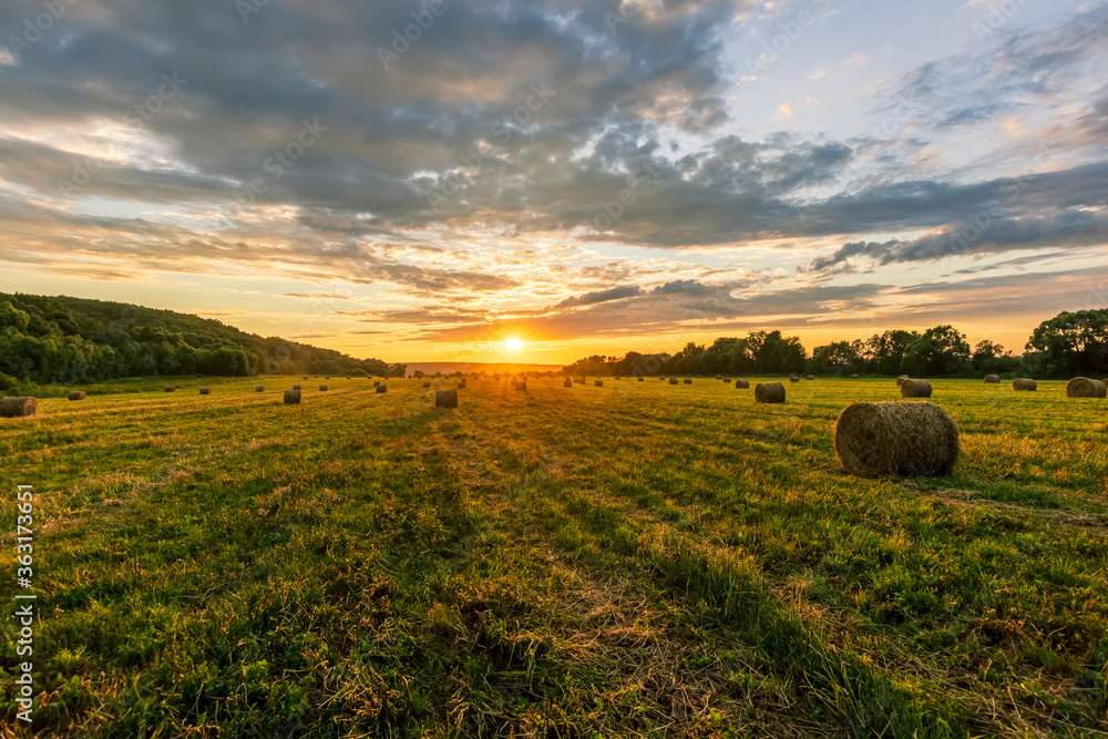 Scenic view at beautiful sunset in a green shiny field with hay stacks, bright cloudy sky , trees and golden sun rays with glow, summer valley landscape