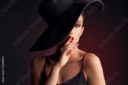 portrait of beautiful tanned girl with professional makeup, red lips, on a burgundy background in a black dress with straps and black hat that covers her eye, she looks at the camera touching her lips